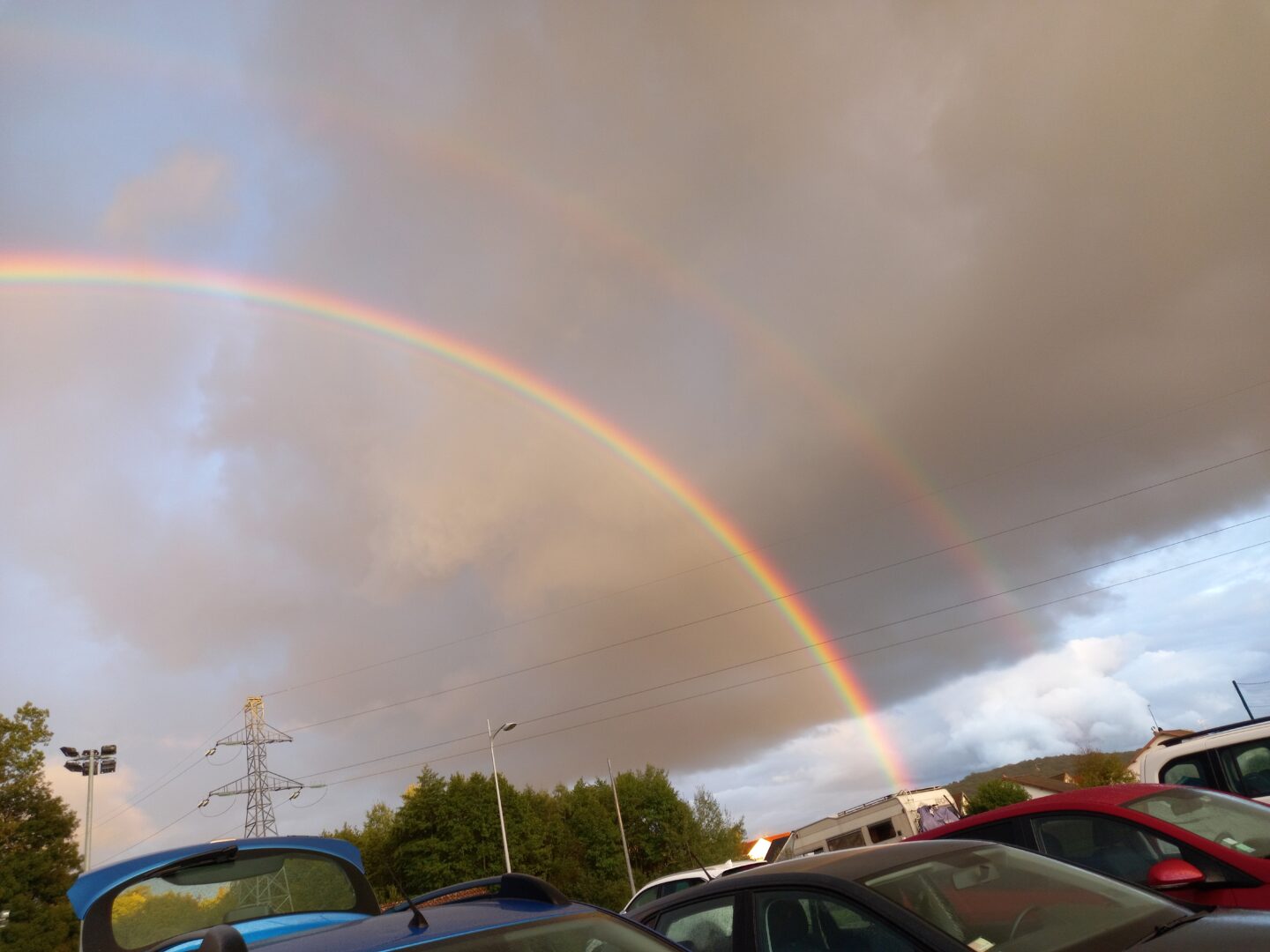 [Justine CM] Double arc-en-ciel pris de dehors qui fait un demi-cercle, vue dégagée. Ciel gris-bleu. Photo en deux parties, 2ème moitié ici.