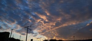 Ciel très nuageux, et ces nuages sont gris, ou orangés avec le soleil couchant derrière. Poteaux électriques.