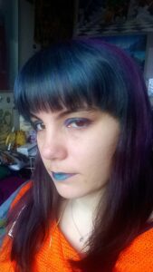 Photo de moi où l'on voir ma frange turquoise et le reste de mes cheveux violets. Fard à paupières, crayon à paupières et rouge à lèvres avec les mêmes teintes. Pull orange.